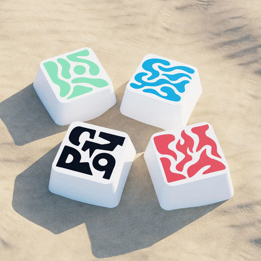 Vue artistique de la collection KCOD Originals Mirage 2025 regroupant quatre keycaps abstraits aux motifs noir, rouge, bleu et vert, posés sur du sable.